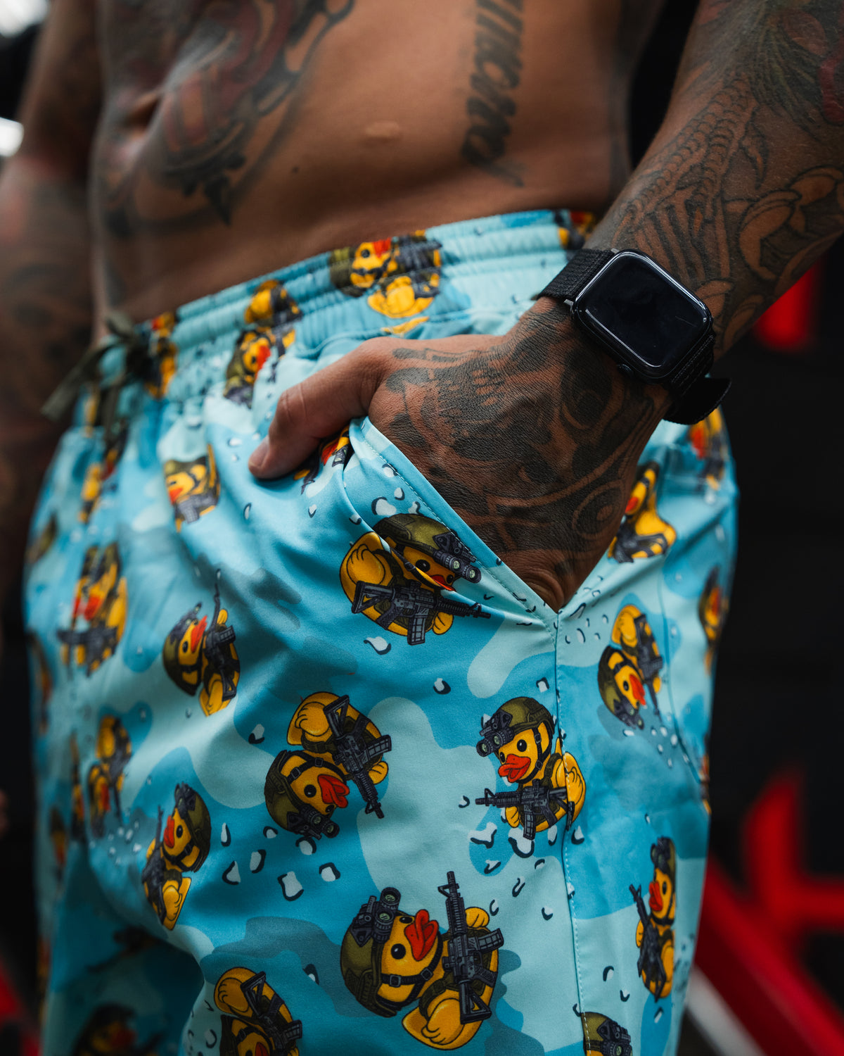 A close-up of a tattooed person wearing Born United Private Label’s Men's Performance Hybrid Shorts 5.5" | TactiQuack, featuring playful rubber duck soldiers. Their hand, with a smartwatch, rests in the pocket of these quick-dry shorts.