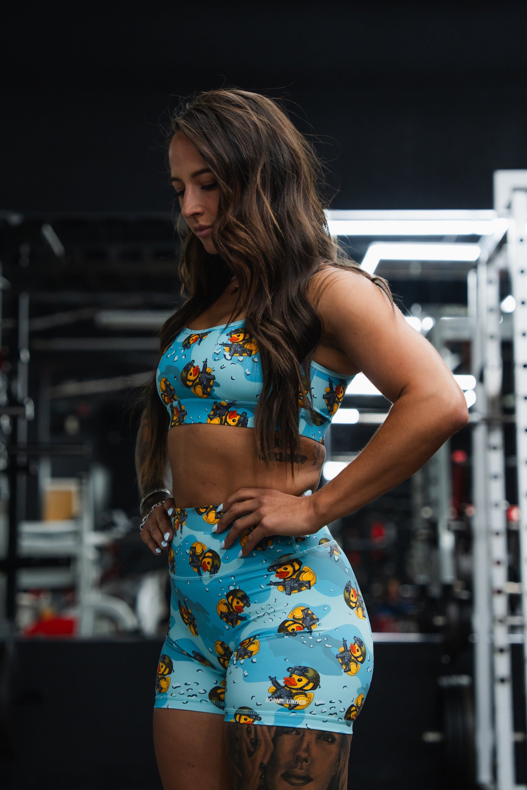 A woman with long brown hair stands in a gym, wearing matching blue Born United Private Label Women's High-Waisted Seamless Front Compression Training Shorts by Tacti-Quack with a bee pattern, looking down with her hands on her hips.