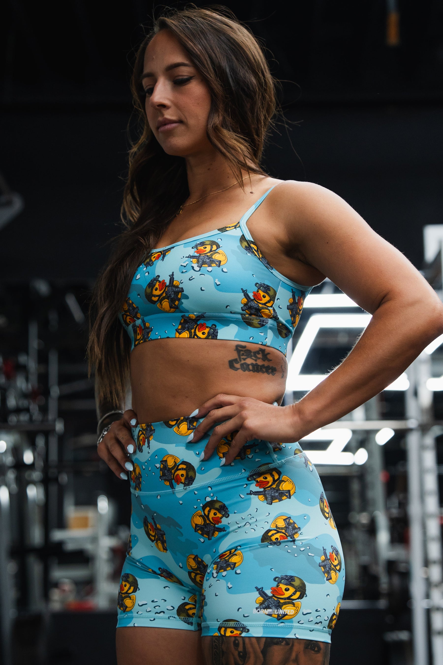 A woman at the gym wears Born United Private Label's Women's High-Waisted Seamless Front Compression Training Shorts in the Tacti-Quack pattern, paired with a matching blue sports bra. Fitness equipment appears in the background.