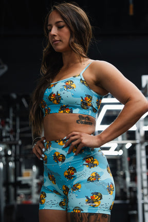 A woman at the gym wears Born United Private Label's Women's High-Waisted Seamless Front Compression Training Shorts in the Tacti-Quack pattern, paired with a matching blue sports bra. Fitness equipment appears in the background.