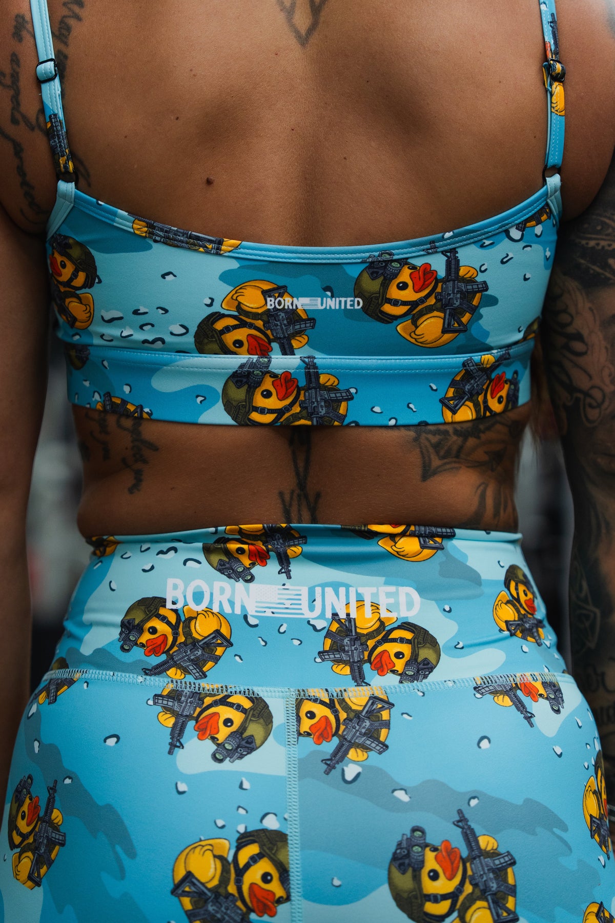 Close-up of a woman's back in Born United Private Label "Tacti-Quack" high-waisted seamless front compression training shorts, sports bra, and leggings, all featuring cartoon military ducks and "BORN UNITED" printed on the bra and leggings.