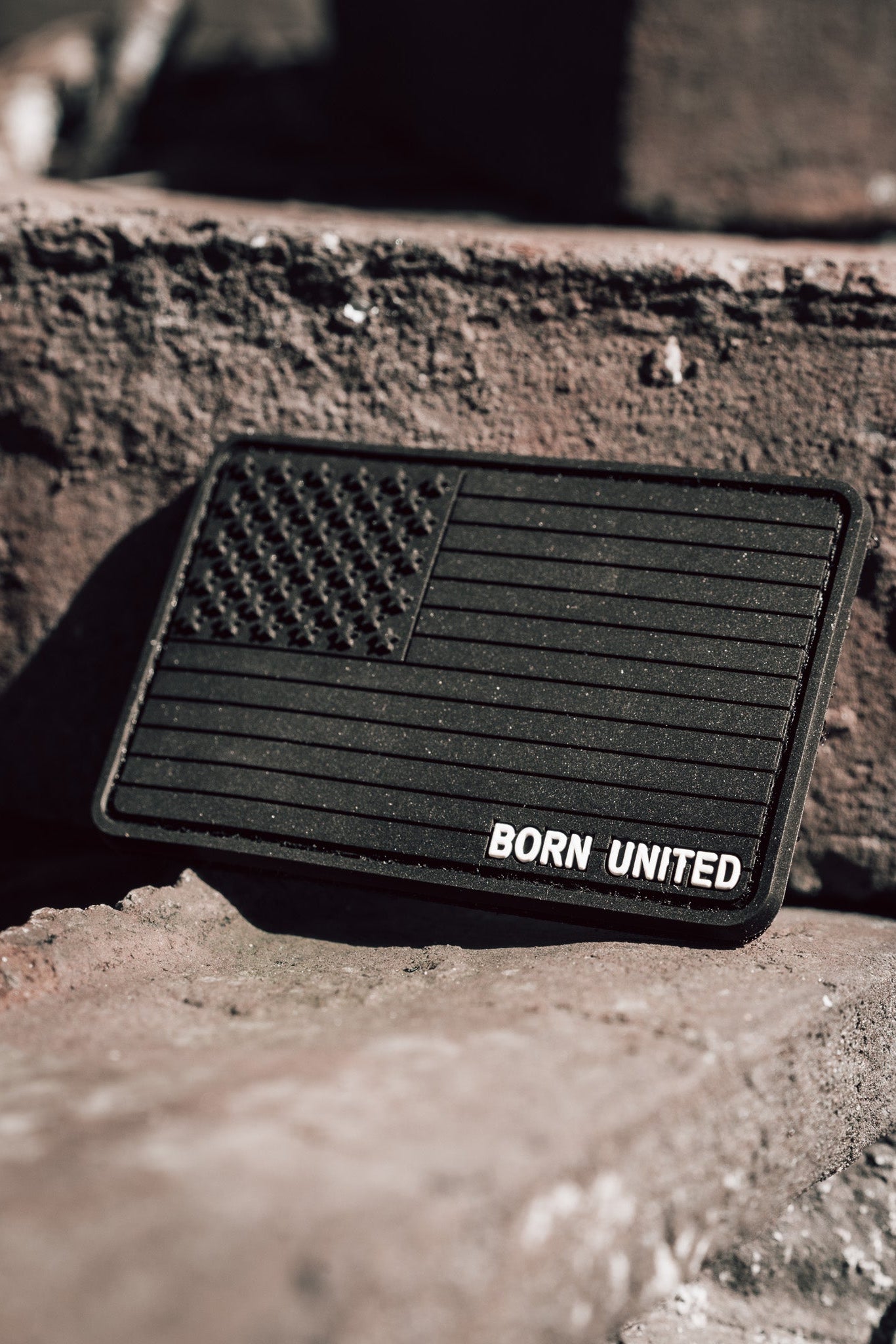 The American Flag PVC Patch - Blackout by Born United Private Label features a monochrome flag with BORN UNITED below, set against a rough brick surface. Angled sunlight highlights its contours, and the hook & loop backing allows for easy attachment.