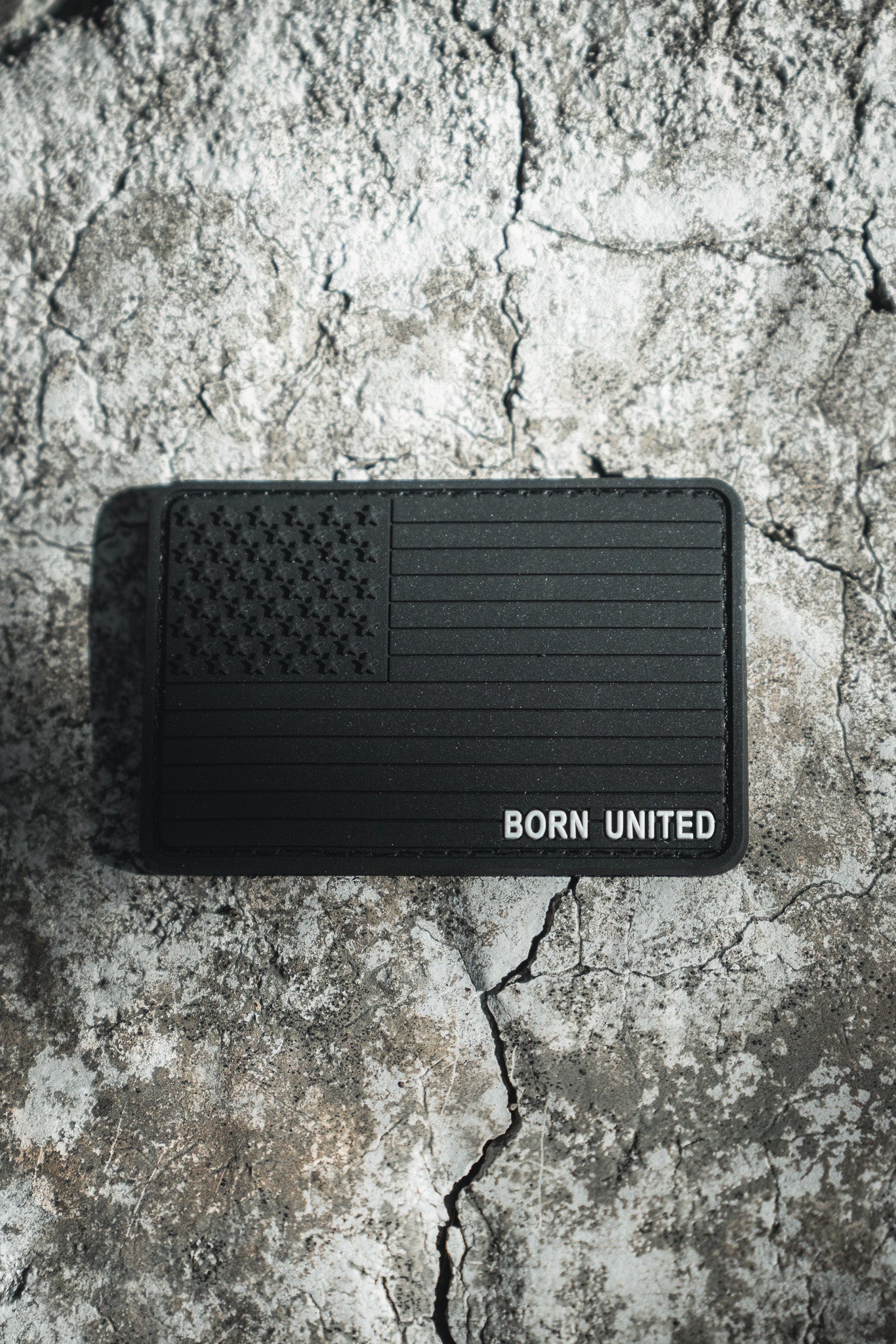 The Born United Private Labels American Flag PVC Patch - Blackout has a textured design featuring the black American flag with BORN UNITED, set against a rugged, stone-like surface. Its hook & loop backing ensures easy attachment, emphasizing its standout design against the rough background.