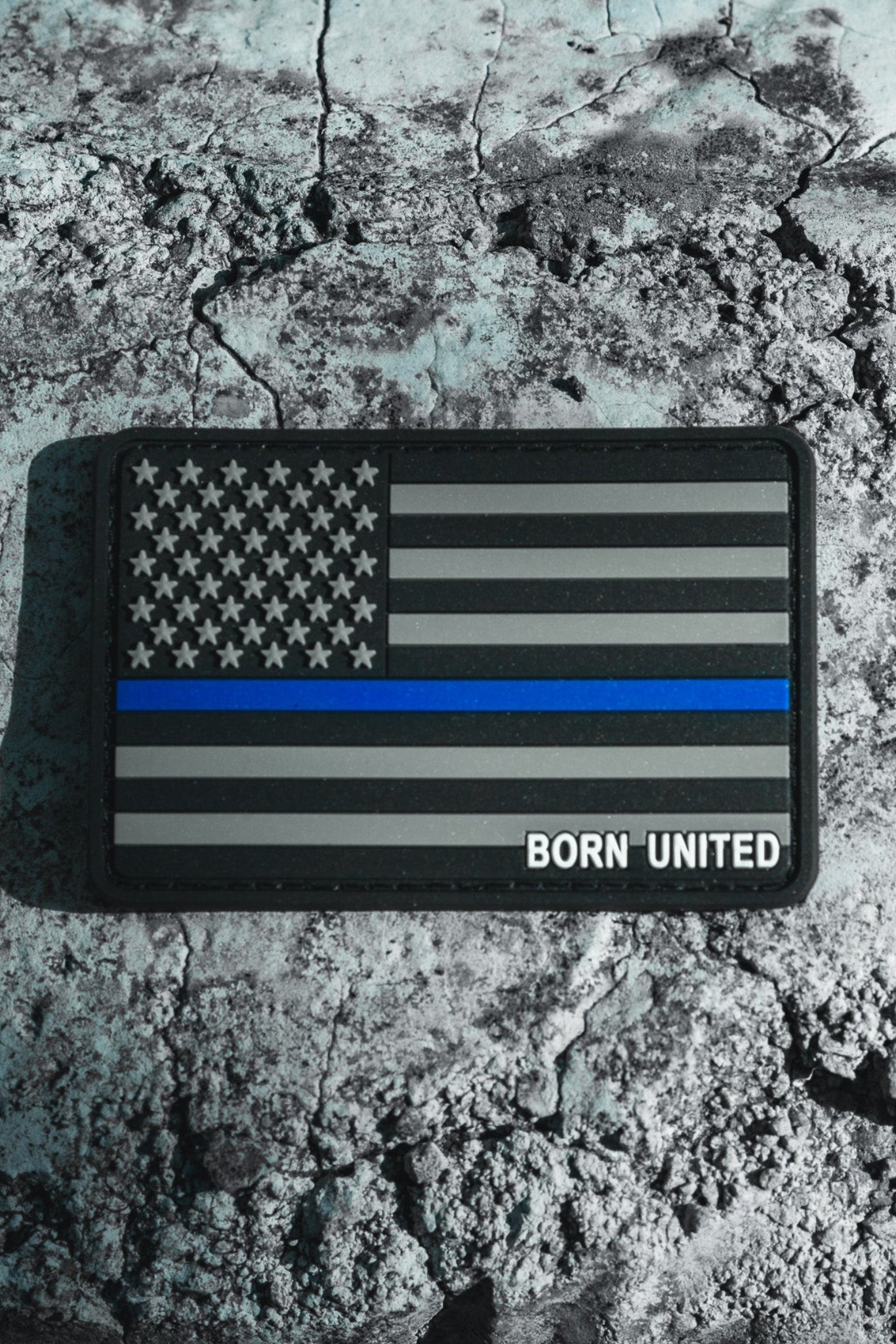 A resilient PVC patch named American Flag PVC Patch - TBL by Born United Private Label, features a grayscale flag with a bold blue stripe symbolizing law enforcement support, resting on cracked earth. Born United is elegantly inscribed in the lower right corner.