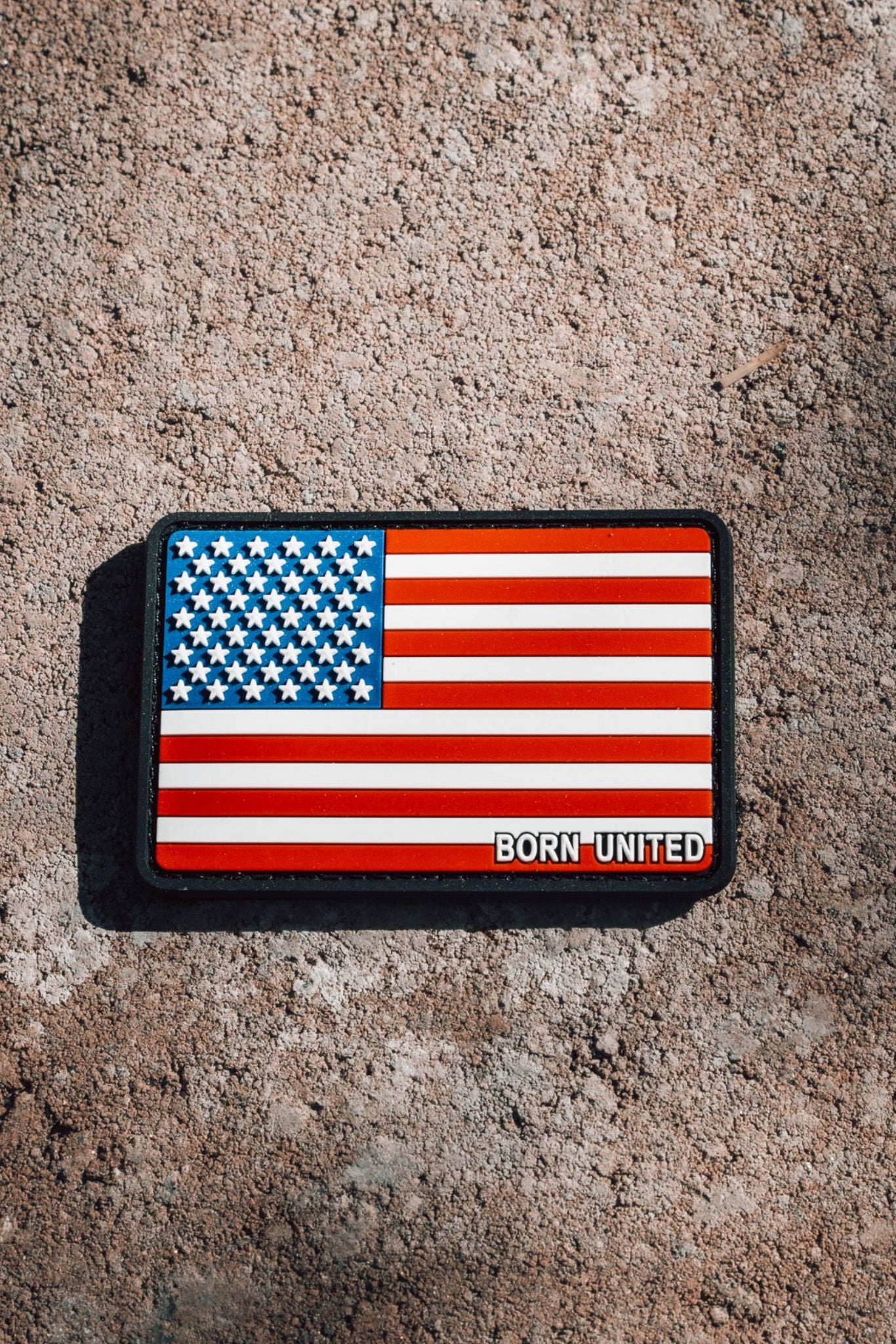 The American Flag PVC Patch by Born United Private Label rests on a rugged cement surface, with Born United boldly displayed. Its waterproof and washable, ensuring durability even in harsh conditions.