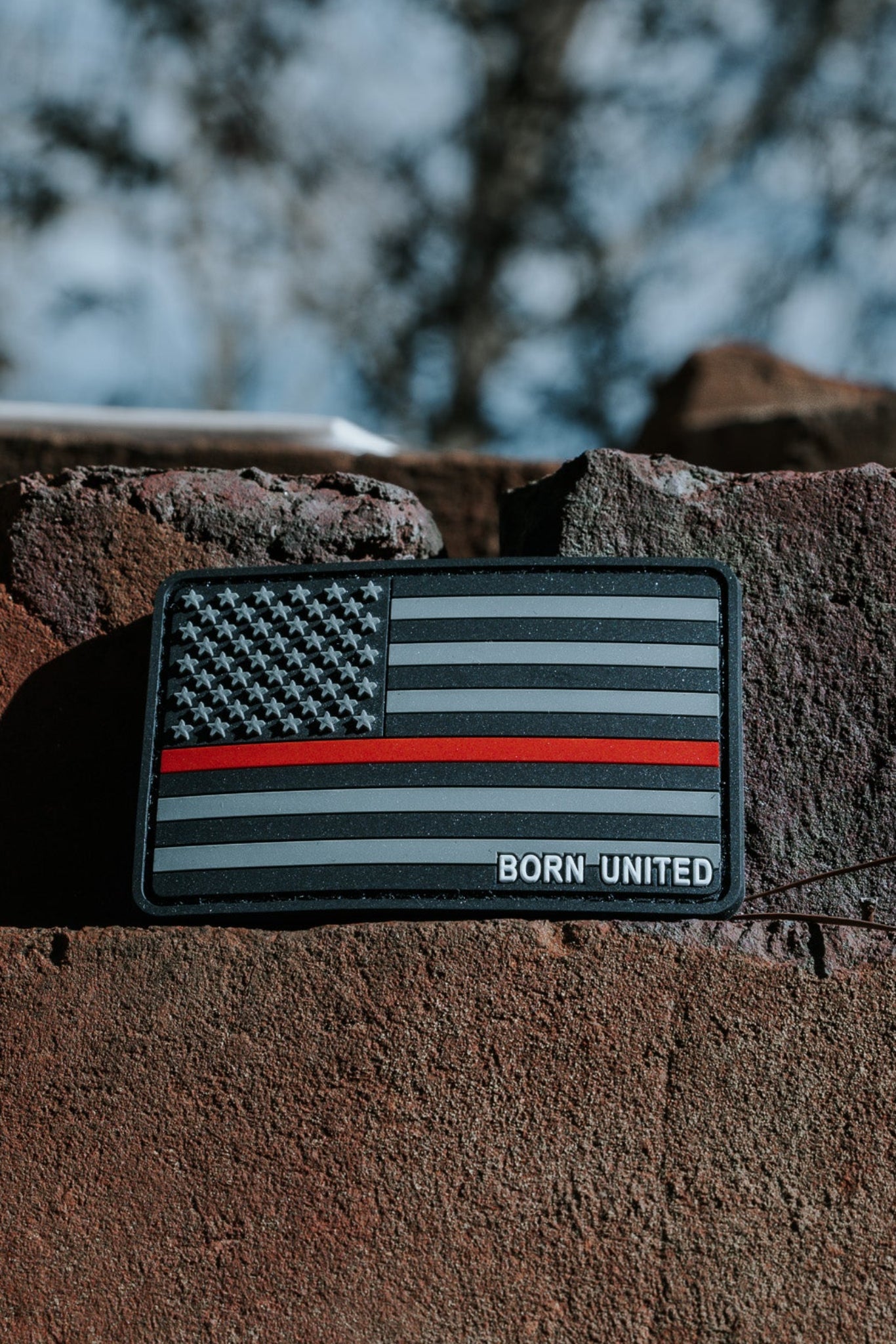 A Born United Private Label PVC patch, featuring a black and gray American flag with a red stripe symbolizing firefighter support, is showcased on a rough brick surface. The words BORN UNITED are visible against a blurred natural background.