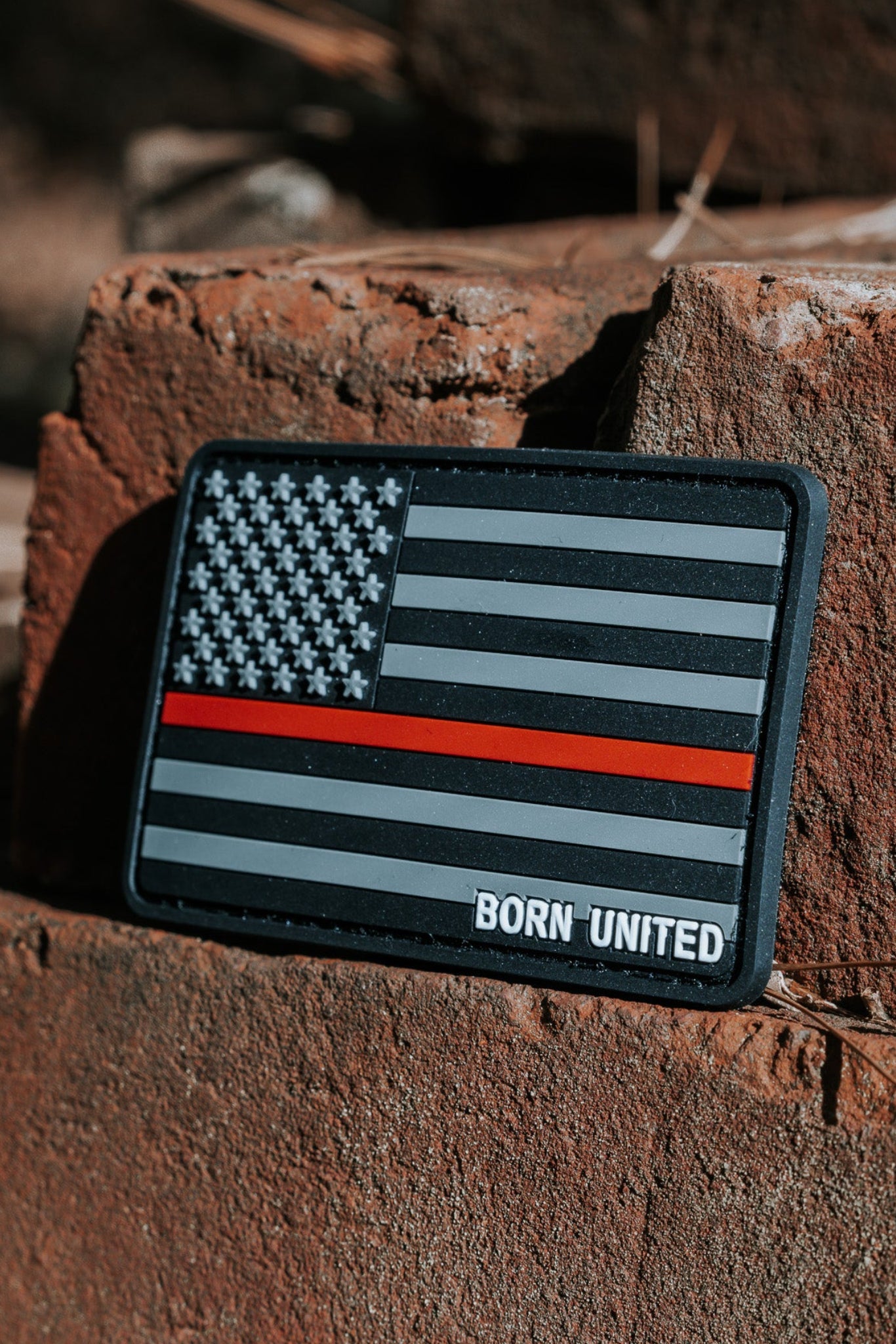 An American Flag PVC Patch - TRL by Born United Private Label, featuring a Thin Red Line and BORN UNITED text below, is displayed on weathered bricks outdoors with natural light casting complex shadows.
