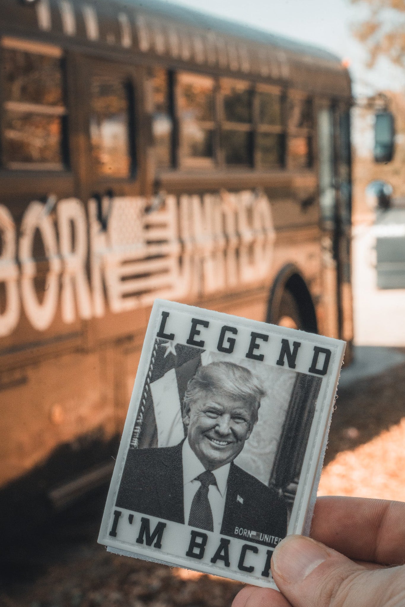 A hand holds the I’m Back PVC Patch from Born United Private Label, showing a smiling man in a suit with "LEGEND IM BACK!", in front of a bus featuring "BORN UNITED," an American flag, and Trump 2024 graphics.