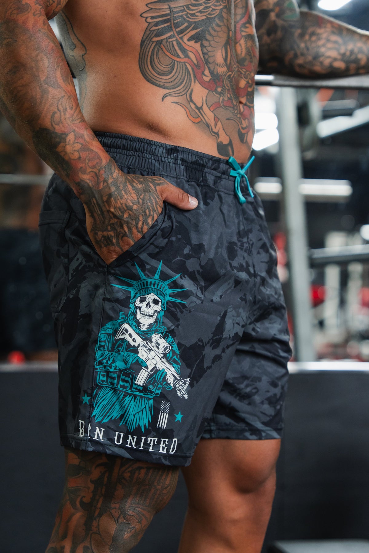 A tattooed, shirtless man stands indoors with one hand in the pocket of Born United Private Label Men's Performance Hybrid Shorts 5.5" | Liberty or Death, featuring quick-drying fabric and a skeleton Statue of Liberty design above "BIN UNITED.