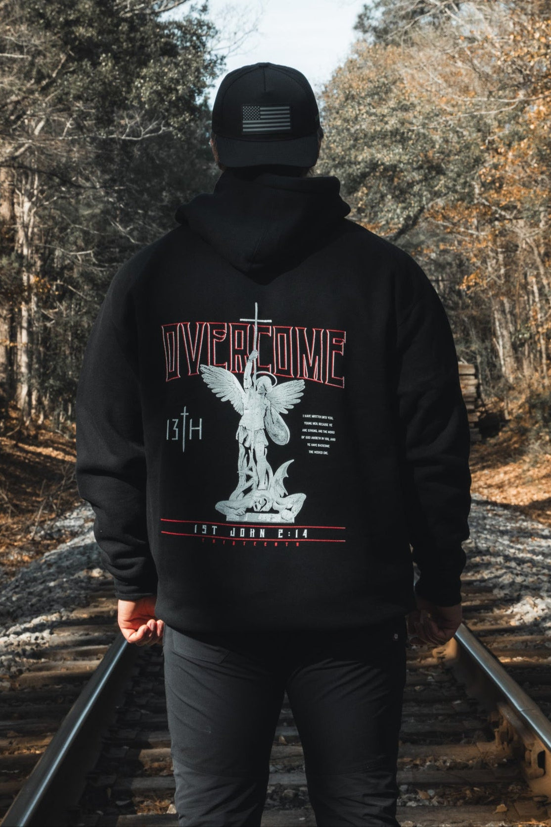 A young man in a black 13th Overcome Premium Hoodie and cap stands on railroad tracks among trees. The hoodie’s back shows an angel statue, "OVERCOME," and faith-inspired motifs referencing the word of God.