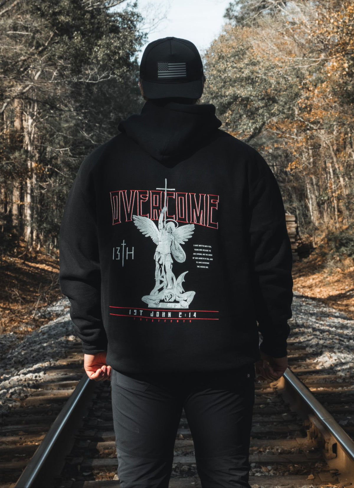 A young man in a black 13th Overcome Premium Hoodie and cap stands on railroad tracks among trees. The hoodie’s back shows an angel statue, "OVERCOME," and faith-inspired motifs referencing the word of God.