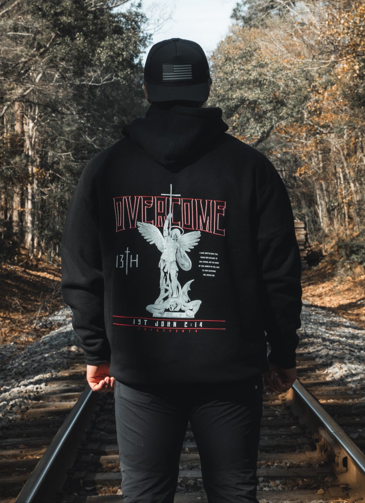 A young man in a black 13th Overcome Premium Hoodie and cap stands on railroad tracks among trees. The hoodie’s back shows an angel statue, "OVERCOME," and faith-inspired motifs referencing the word of God.