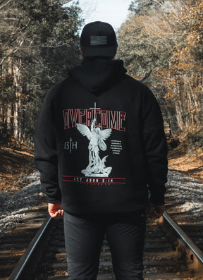 A young man in a black 13th Overcome Premium Hoodie and cap stands on railroad tracks among trees. The hoodie’s back shows an angel statue, "OVERCOME," and faith-inspired motifs referencing the word of God.