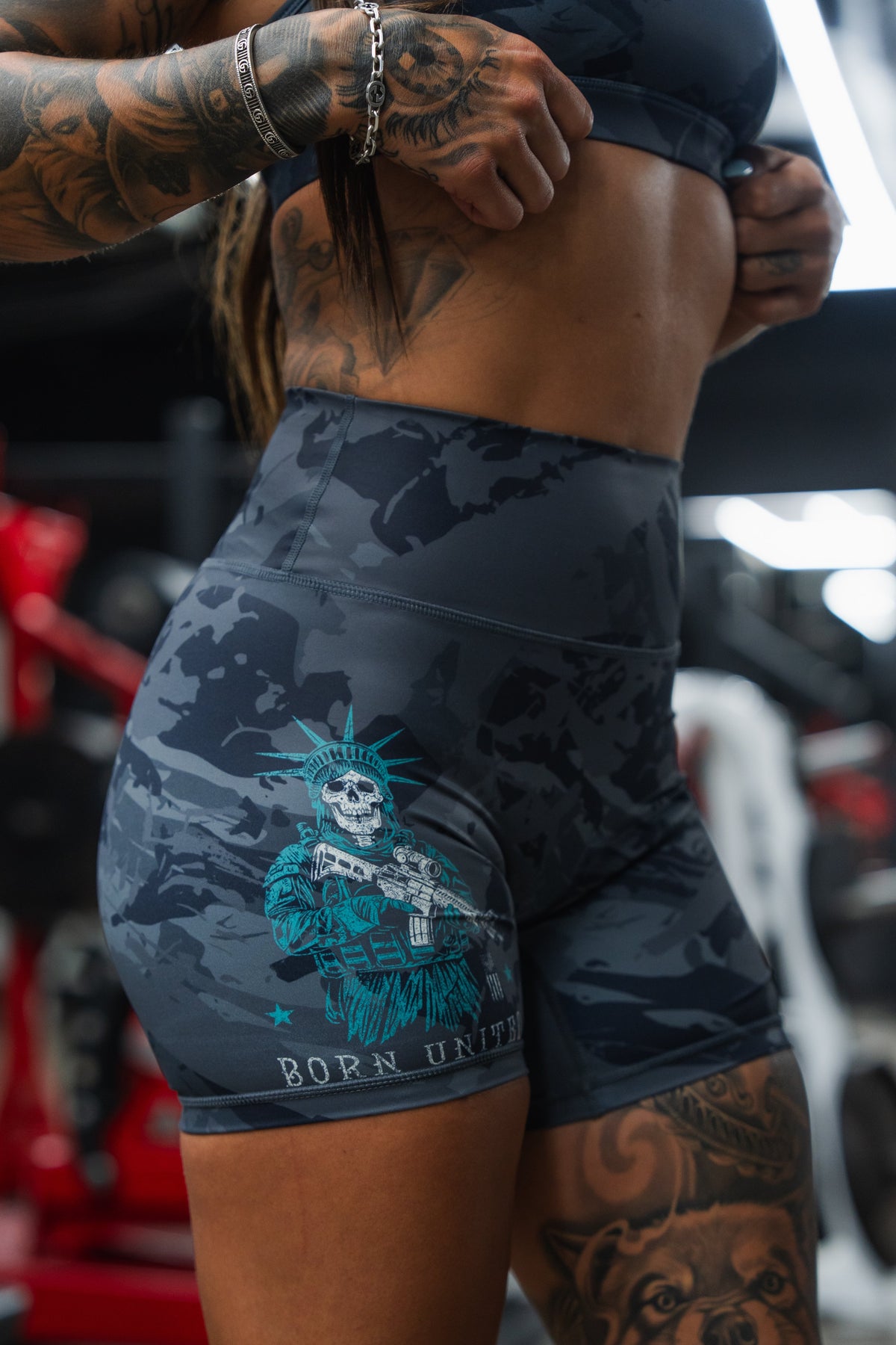 A tattooed individual stands in a gym wearing Born United Private Label Women's High-Waisted Seamless Front Compression Training Shorts, featuring a skeleton with a rifle and "Liberty or Death" graphic. Blurred gym equipment is visible behind them.