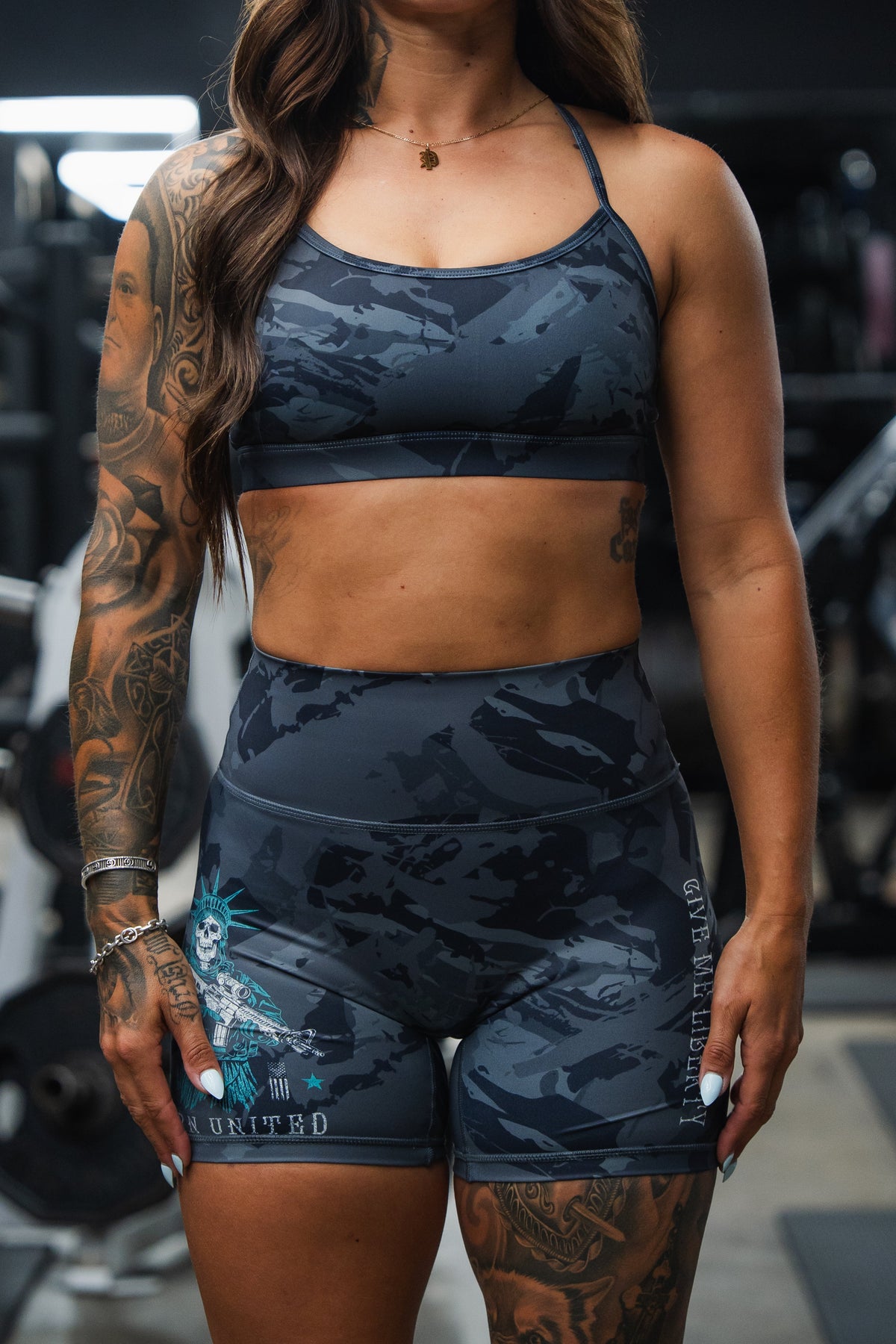 A woman with long wavy hair and tattooed arms stands in a gym, wearing Born United Private Label’s Women's High-Waisted Seamless Front Compression Training Shorts | Liberty or Death—ideal for intense, breathable workout sessions.