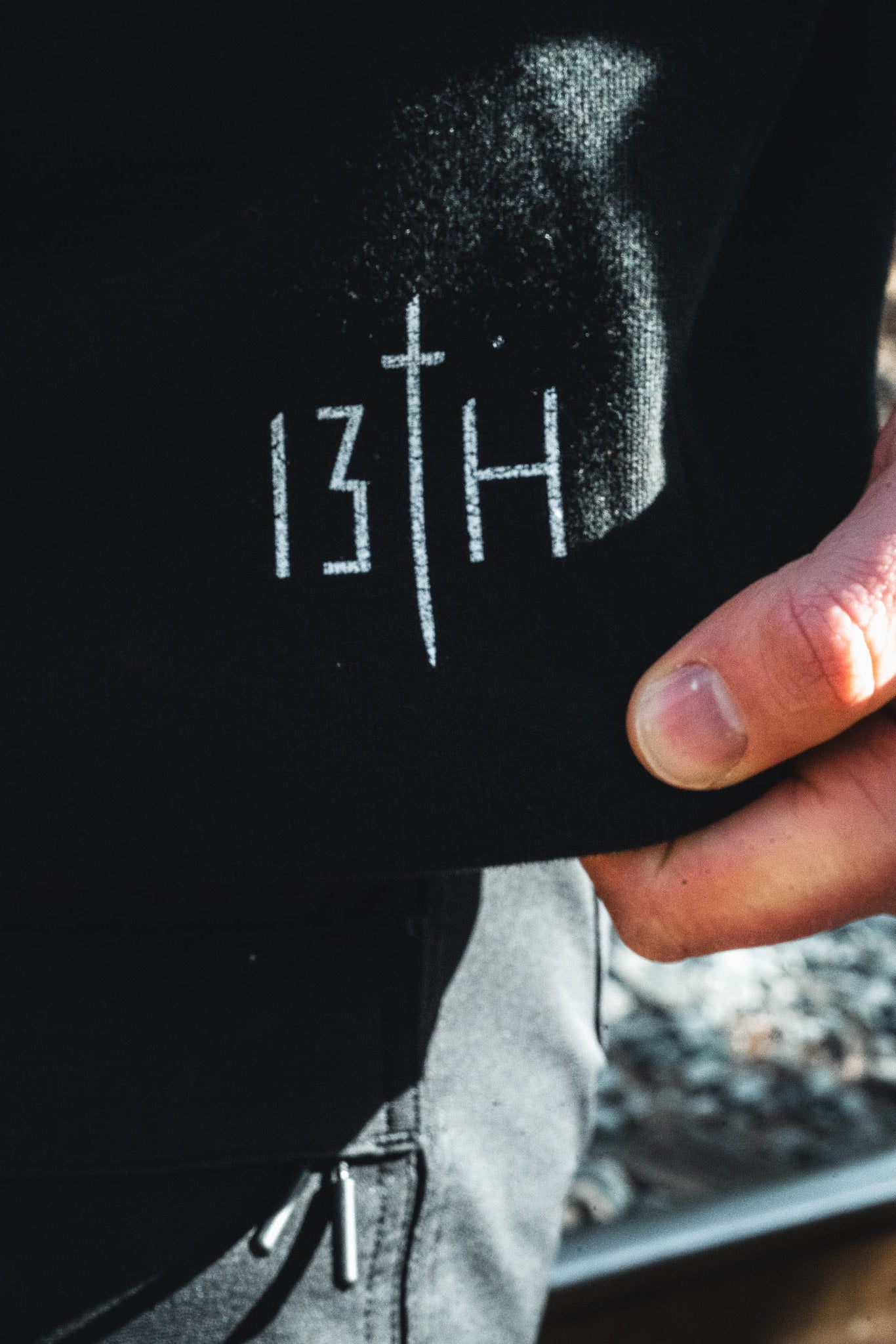A close-up of a young man's hand holding the edge of a black Overcome Premium Hoodie by 13th, featuring white text and a vertical line reading "13TH." The slightly blurred background reveals outdoor scenery.