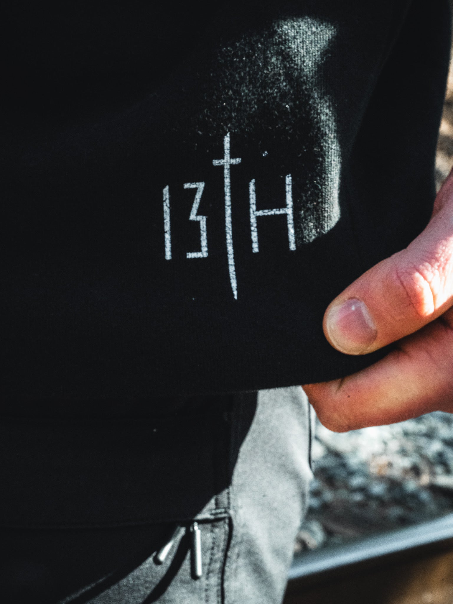 A close-up of a young man's hand holding the edge of a black Overcome Premium Hoodie by 13th, featuring white text and a vertical line reading "13TH." The slightly blurred background reveals outdoor scenery.