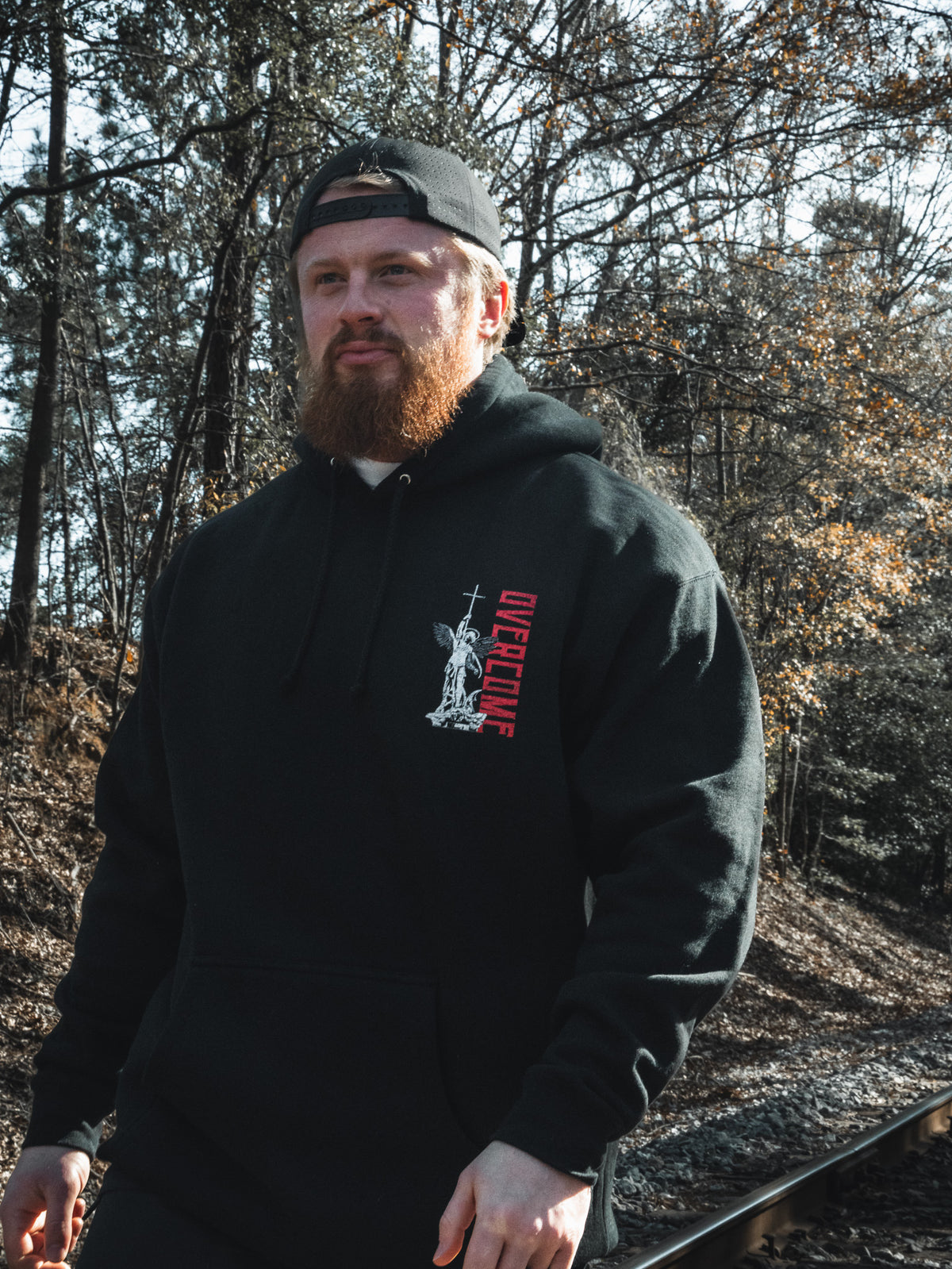 A man with a red beard, wearing the Overcome Premium Hoodie by 13th and a backward cap, walks beside railroad tracks through sunlit woods—a quiet moment reflecting youthful spiritual searching.