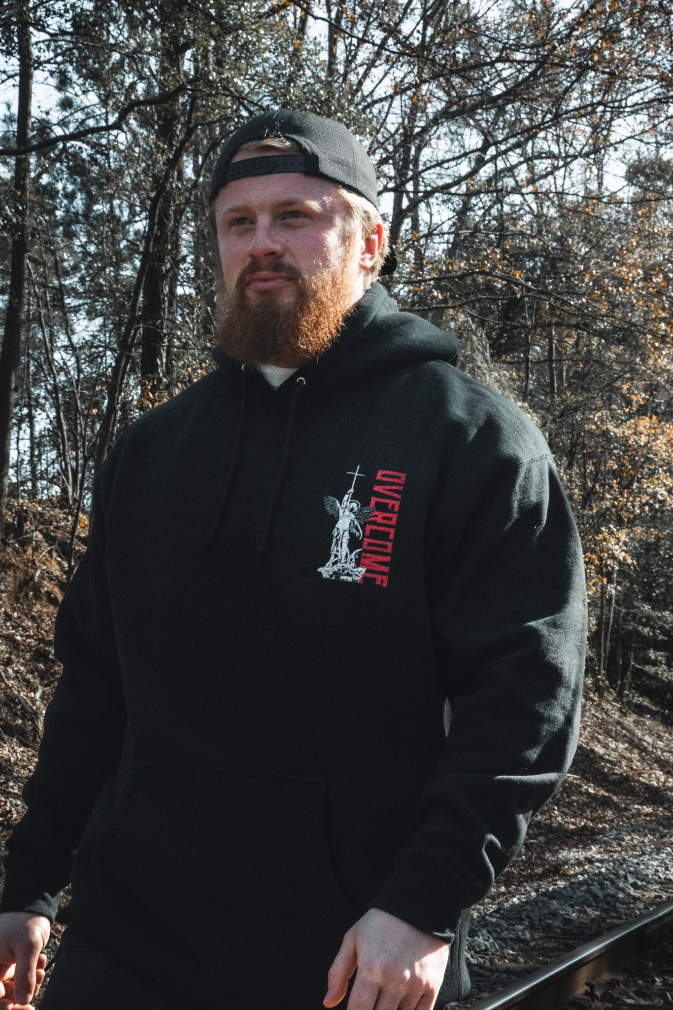 A man with a red beard, wearing the Overcome Premium Hoodie by 13th and a backward cap, walks beside railroad tracks through sunlit woods—a quiet moment reflecting youthful spiritual searching.