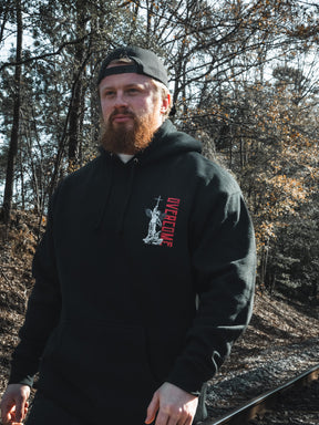 A man with a red beard, wearing the Overcome Premium Hoodie by 13th and a backward cap, walks beside railroad tracks through sunlit woods—a quiet moment reflecting youthful spiritual searching.