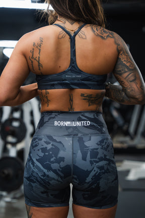 A tattooed woman in a blue sports bra and Born United Private Label Women's High-Waisted Seamless Front Compression Training Shorts (Liberty or Death) stands in a gym with "BORN UNITED" on the waistband, gym equipment behind her.