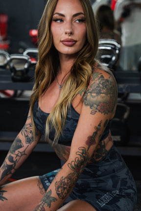 A woman with long wavy hair and tattoos sits in a gym, wearing a dark sports bra and Born United Private Label’s Women's High-Waisted Seamless Front Compression Training Shorts | Liberty or Death. Kettlebells are visible in the background.