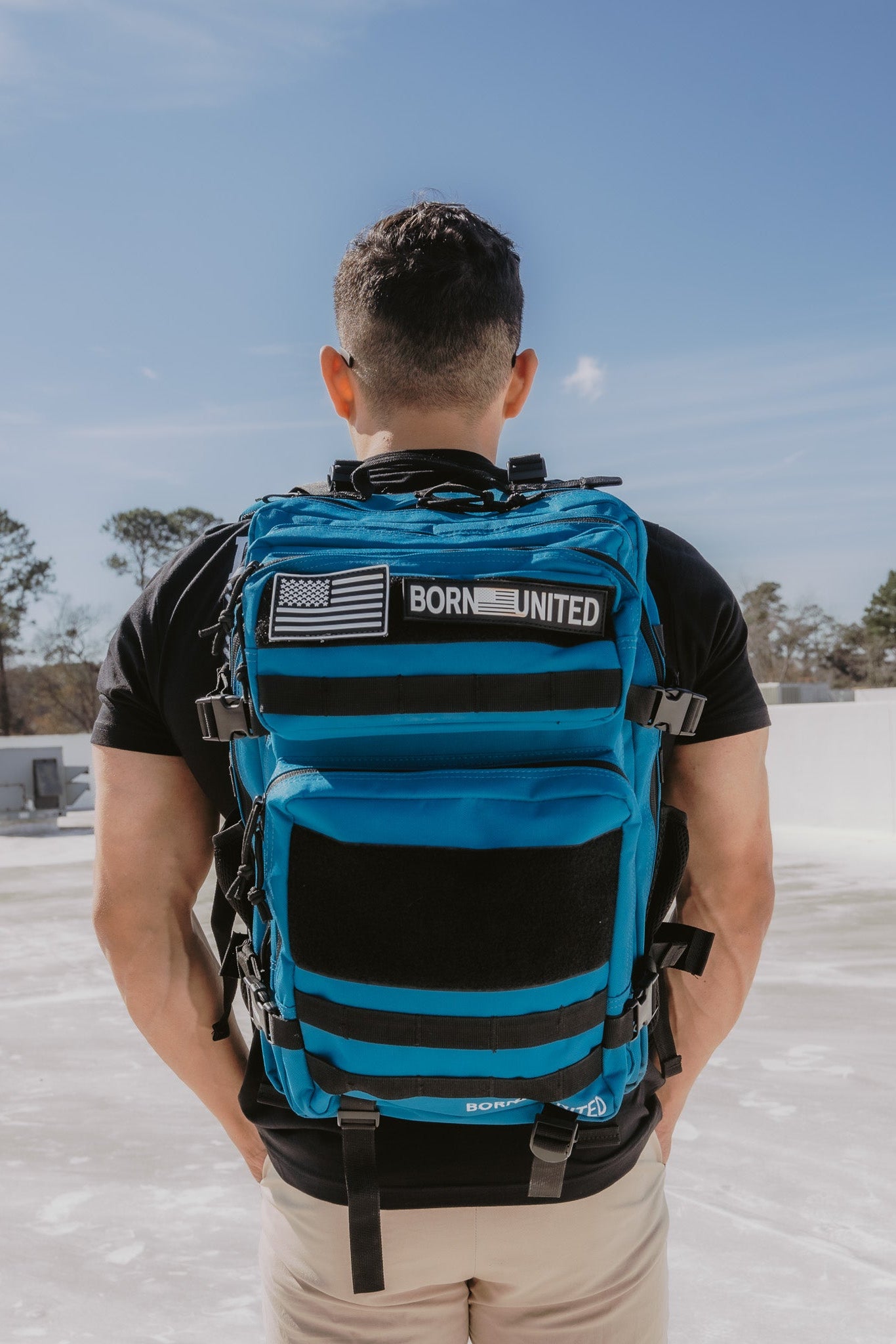 A person with short hair stands outside, back turned, wearing a black shirt and beige pants. They carry the 45L Electric Blue Premium Tac-Pac by Born United Private Label, featuring black straps and an American flag patch. The sky is clear, with trees visible in the background.