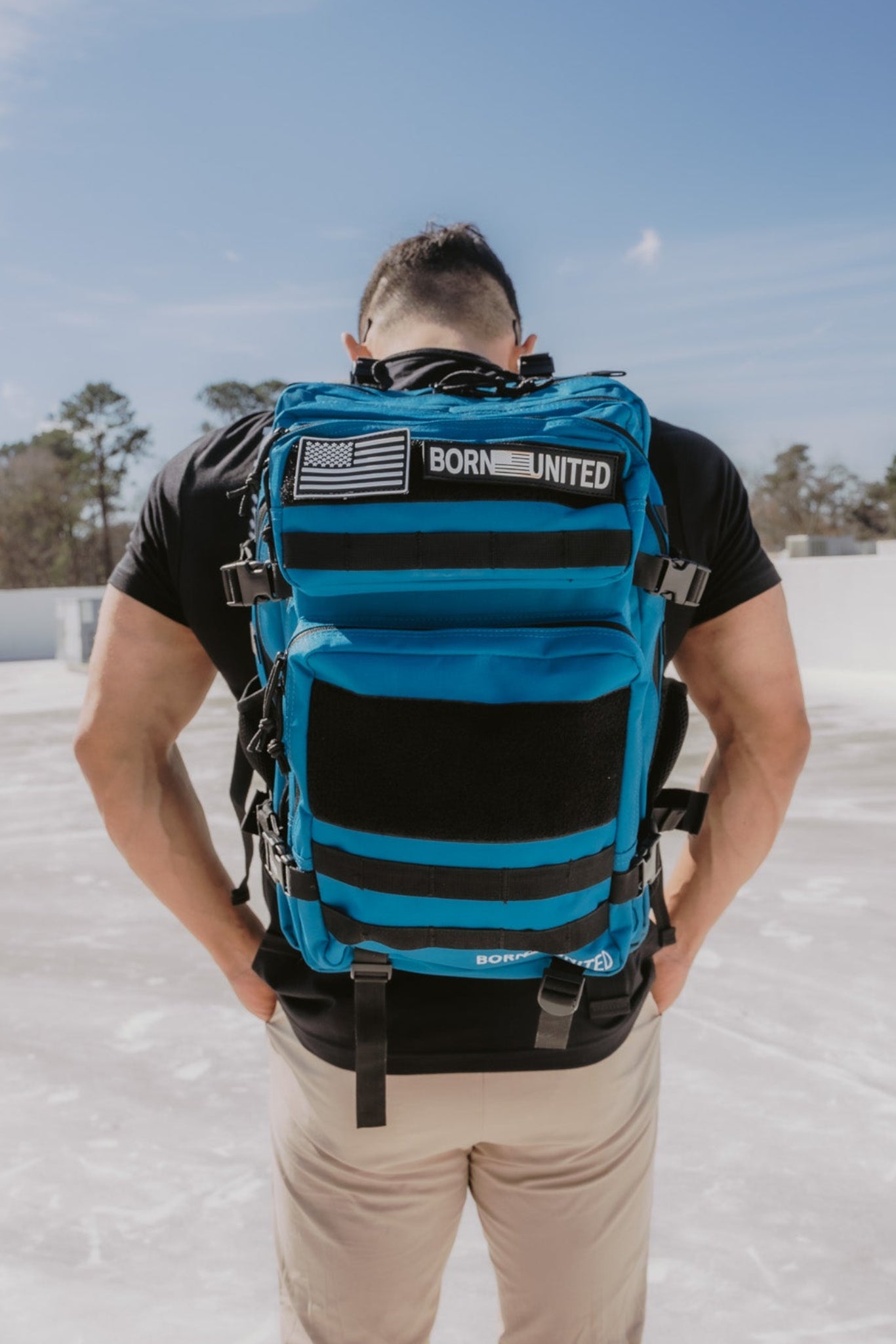 Standing outside on a sunny day, a person in a black shirt and beige pants is seen from behind wearing the Electric Blue Premium Tac-Pac - 45L by Born United Private Label. The serene backdrop features trees and a clear blue sky.