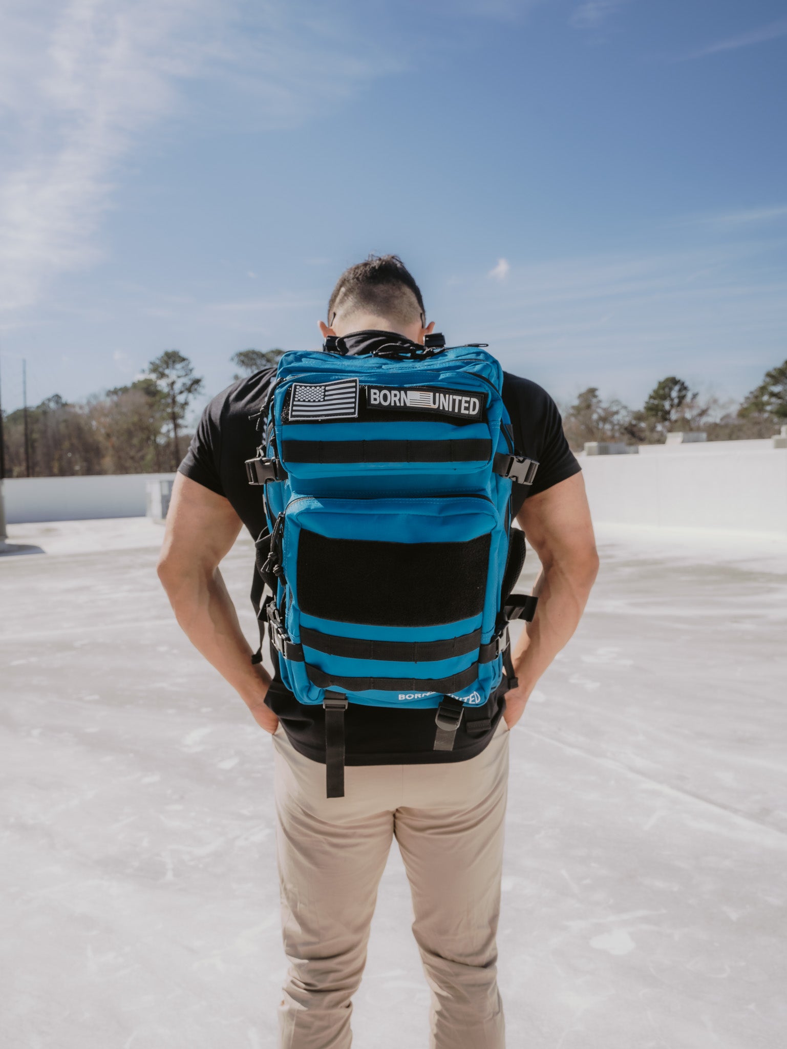 A person stands outdoors on a sunny day, wearing a black shirt and beige pants, with a large Premium 45L Tac-Pac Ultimate Bundle by Born United featuring patches, including an American flag. The background shows a clear sky and distant trees.