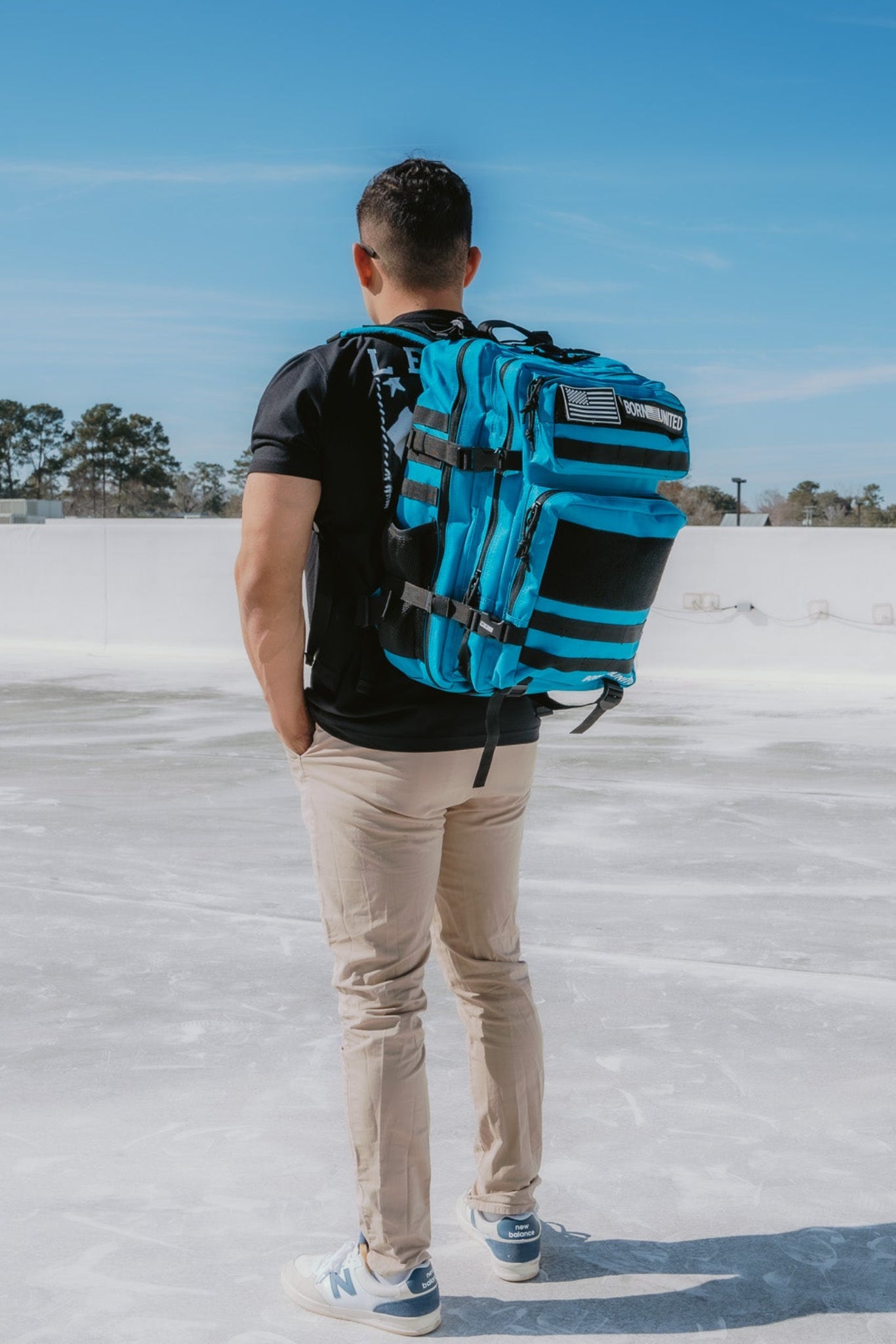 With their back to the camera, a person is outdoors in a black t-shirt, beige pants, and white sneakers. They carry a Born United Private Label Electric Blue Premium Tac-Pac - 45L backpack that contrasts vividly against the clear sky while standing on a paved surface with distant trees.