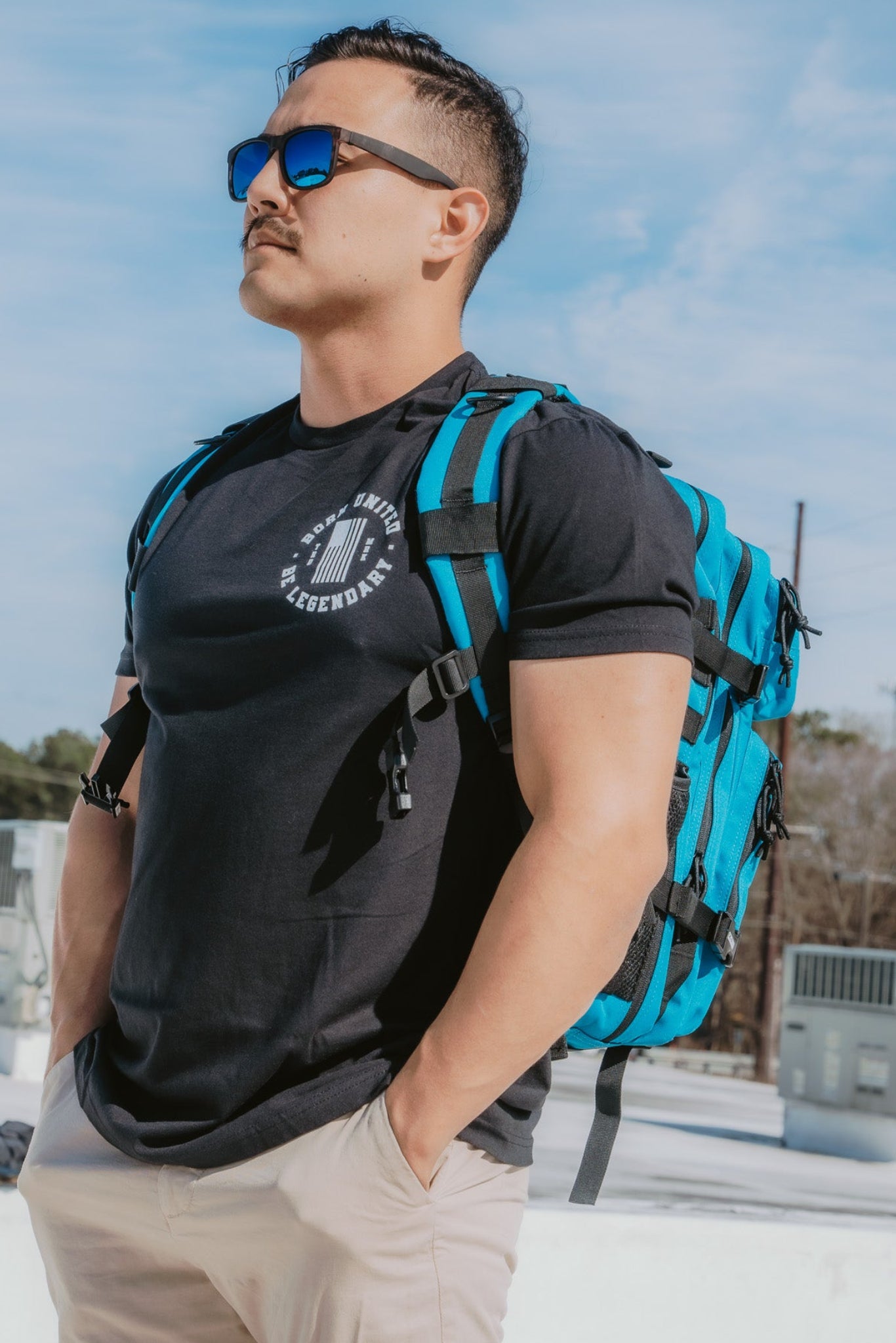 A man with short hair, wearing electric sunglasses and a black T-shirt, pairs his look with beige pants and the Electric Blue Premium Tac-Pac - 45L by Born United Private Label. He stands outdoors confidently with hands in pockets, as the sky and buildings set the scene behind him.
