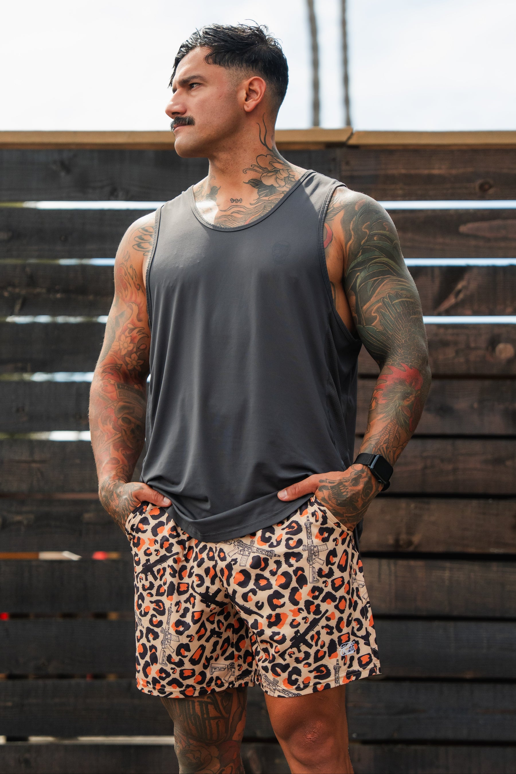A tattooed man in a gray tank top and Tango Charlie Apparel Men's Performance Training Shorts | 5.5"–Loaded Leopard stands outdoors, hands in pockets, looking to the side by a dark wooden fence.