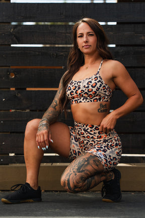 A woman with long brown hair and tattoos kneels confidently outdoors in front of a dark wooden fence, wearing Tango Charlie Apparel’s Women's High-Waisted Seamless Front Compression Training Shorts in Loaded Leopard print.