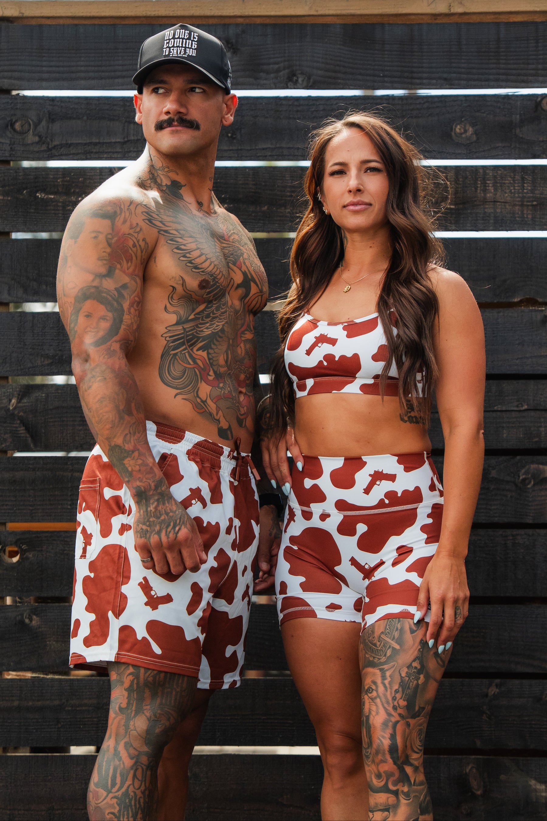 A man in a black hat and red-and-white cow print swim trunks poses confidently with a woman wearing Tango Charlie Apparel’s Women's High-Waisted Seamless Front Compression Training Shorts in Moo Moo Pew, both standing in front of a wooden fence.