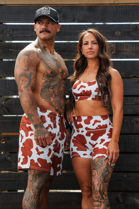 A man in a black hat and red-and-white cow print swim trunks poses confidently with a woman wearing Tango Charlie Apparel’s Women's High-Waisted Seamless Front Compression Training Shorts in Moo Moo Pew, both standing in front of a wooden fence.