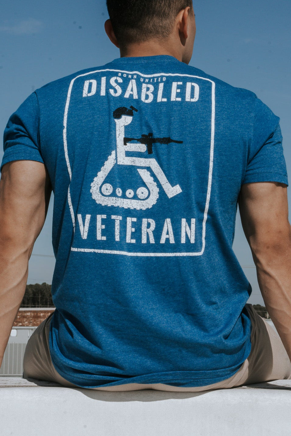 A disgruntled person sits on a ledge against a clear blue sky, wearing the Disabled Vet - Blue T-shirt by Born United Private Label, featuring a graphic of a wheelchair and the text Disabled Veteran.