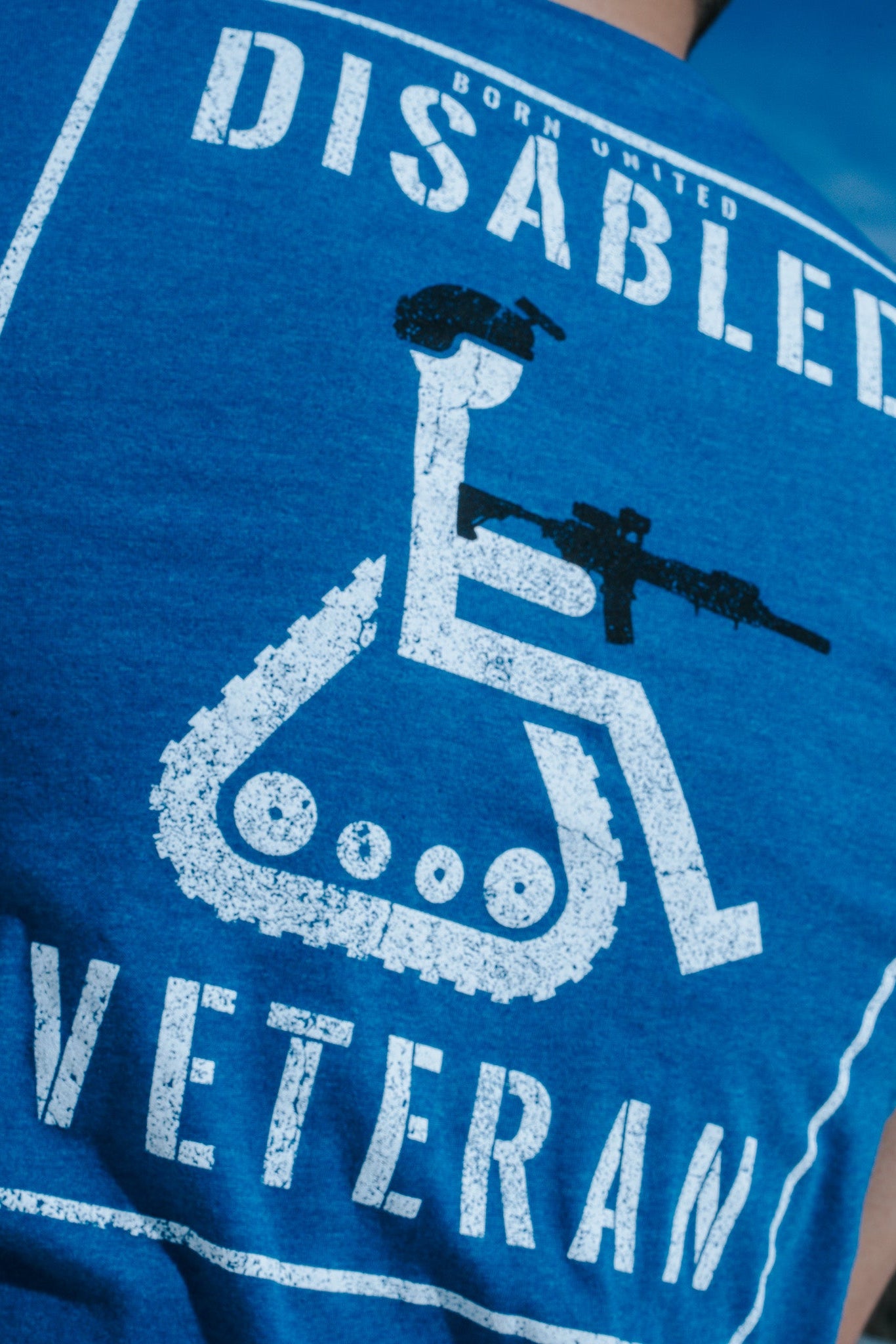 Back view of the Born United Private Label Disabled Vet - Blue T-shirt, featuring a wheelchair symbol with a helmet and rifle, along with Disabled Veteran text. The wearers disgruntled expression complements the outdoor setting against a tranquil blue sky.