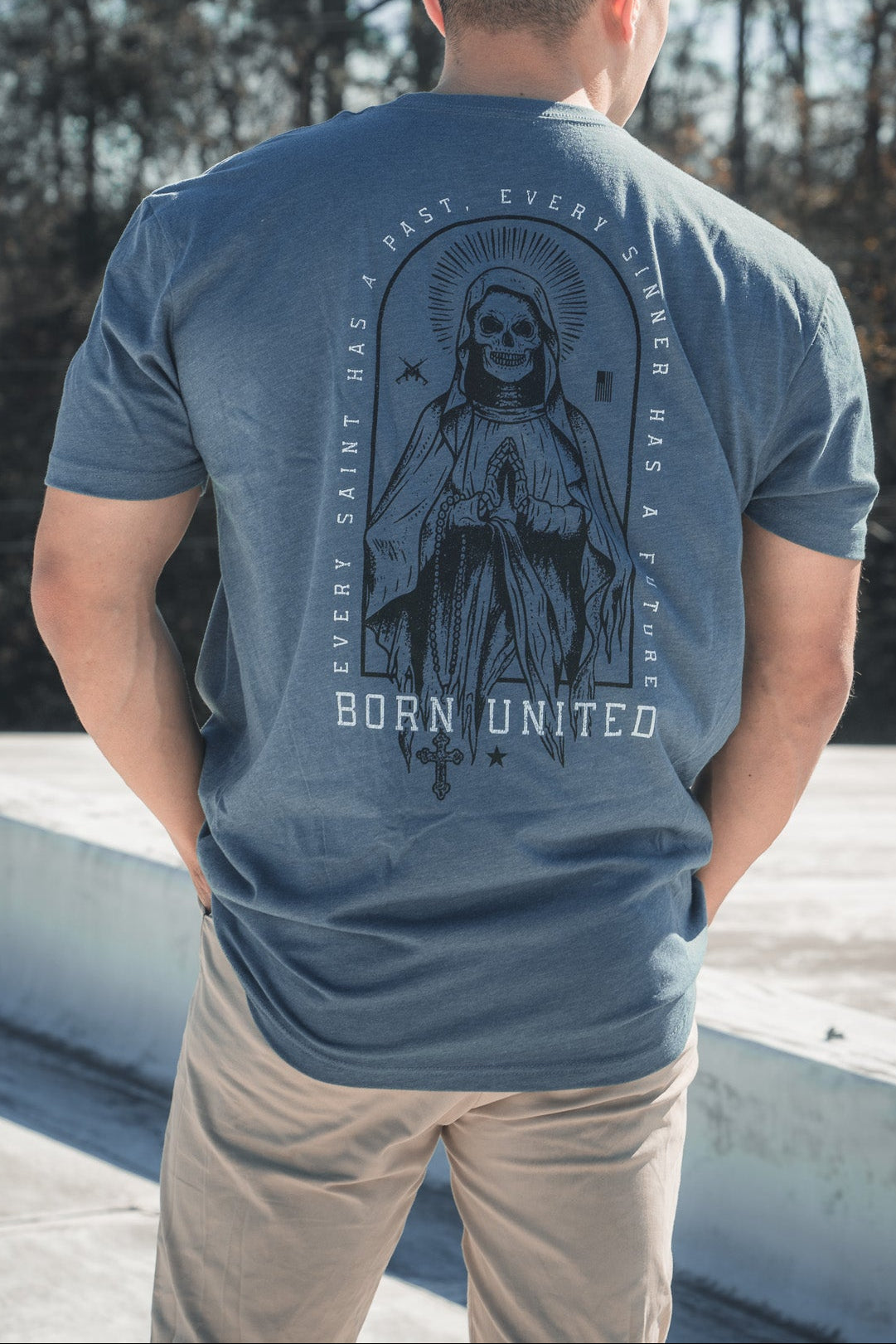 A man outdoors on a rooftop faces away, wearing the Saints And Sinners Tee- Blue by Born United Private Label. The shirt showcases a hooded figure with the text Every Saint Has a Past, Every Sinner Has a Future.