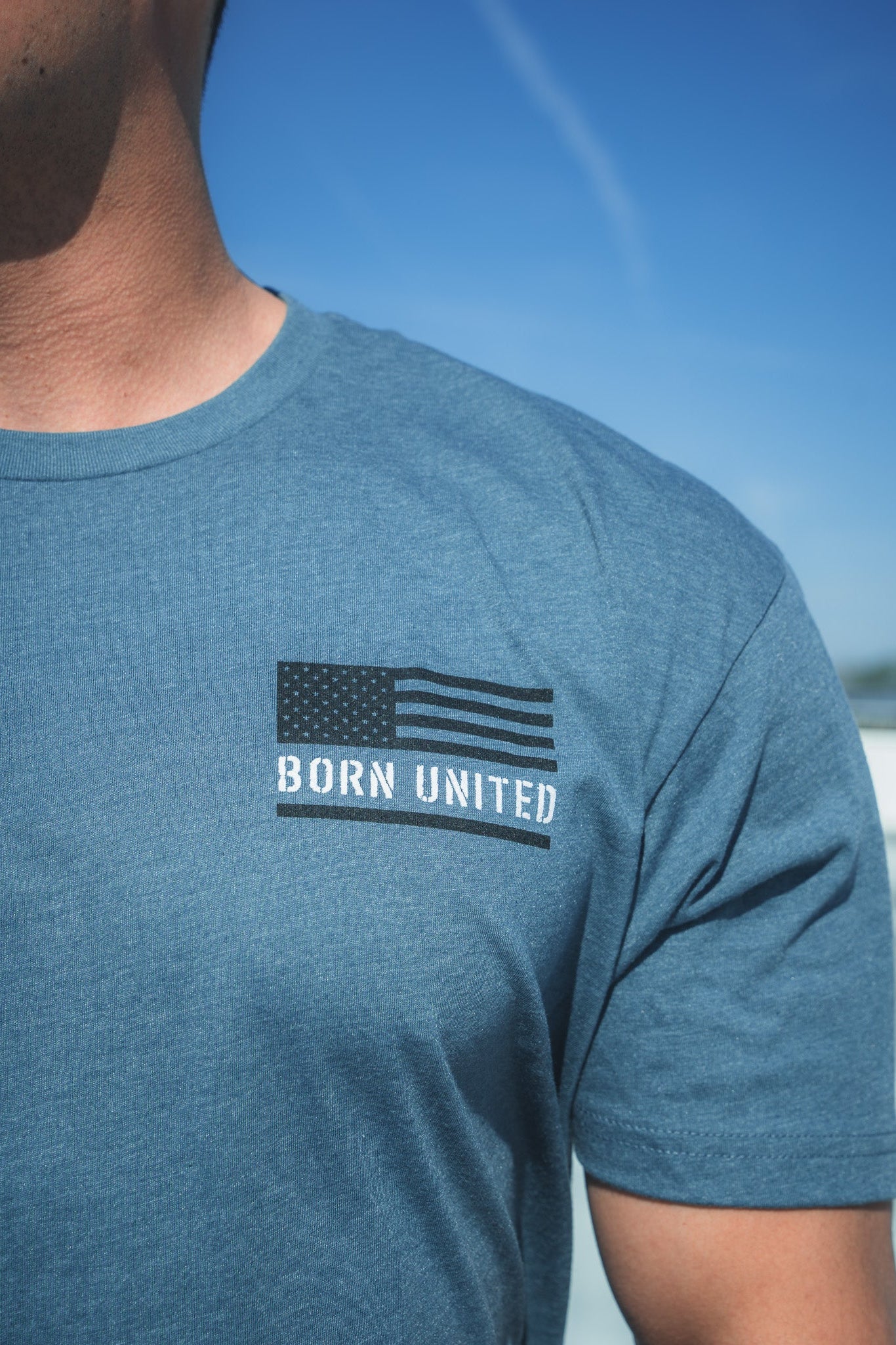 A person wearing the Saints And Sinners Tee- Blue by Born United Private Label, featuring a black and white U.S. flag with BORN UNITED on the chest, stands against a clear blue sky, seemingly pondering the future. Only their upper torso is visible.
