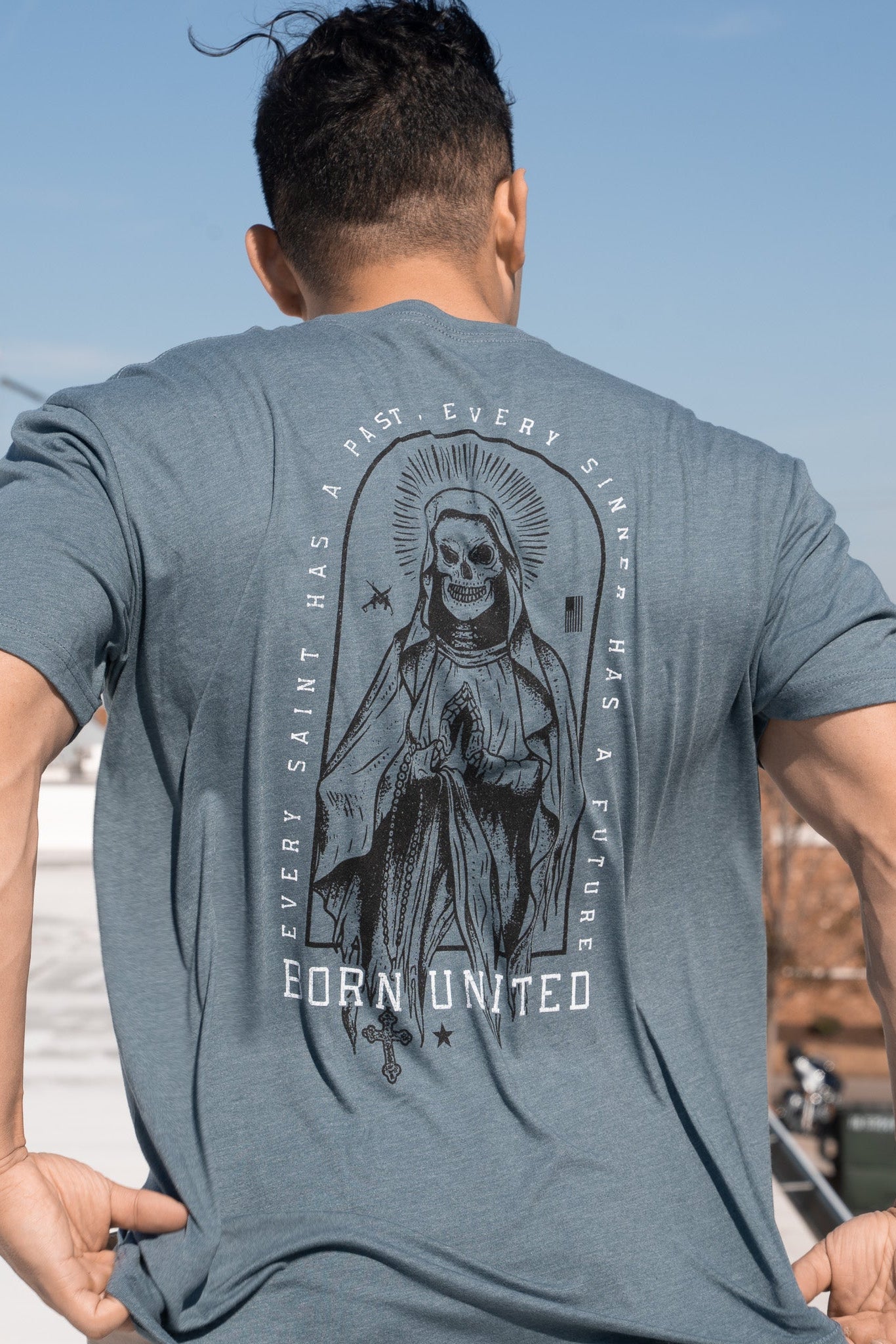 A person is wearing the Saints And Sinners Tee- Blue by Born United Private Label, featuring a skeletal saint in a robe with a halo and prayer hands. The text reads, Every saint has a past, every sinner has a future, with Born United below.