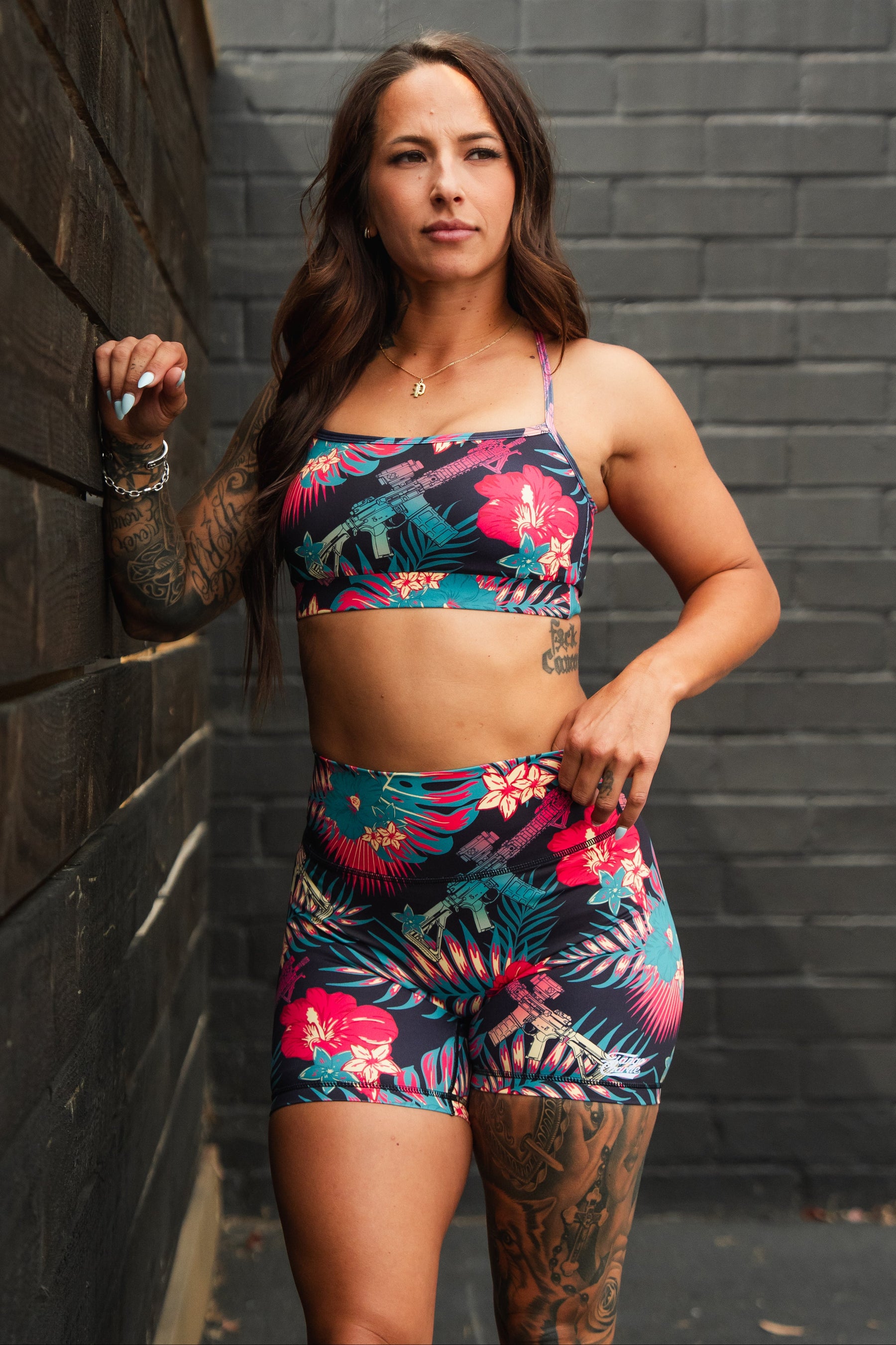 A woman with long brown hair and tattoos wears the Cross Back Sports Bra | Tropical Assassin by Tango Charlie Apparel, standing outdoors near a brick wall and wooden fence, looking confidently into the distance.