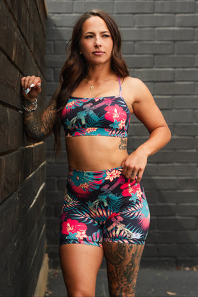 A woman with long brown hair and tattoos poses confidently outdoors, wearing Tango Charlie Apparel's Women's High-Waisted Seamless Front Compression Training Shorts in Tropical Assassin, leaning against a wooden wall by a gray brick background.