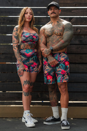 A tattooed woman and man stand side by side in matching tropical floral outfits. She wears Tango Charlie Apparel’s Women's High-Waisted Seamless Front Compression Training Shorts | Tropical Assassin with a crop top, while he sports breathable workout shorts.