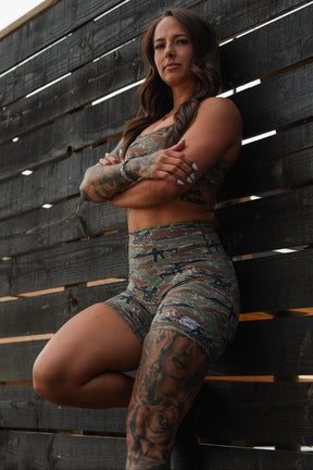 Athletic woman with long brown hair and tattoos wears the Straight Strap Sports Bra | Snipes & Stripes by Tango Charlie Apparel, confidently leaning against a dark wooden fence with arms crossed.