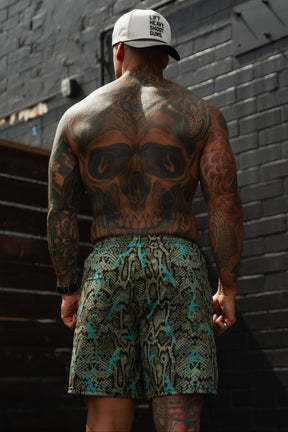 A muscular man in Tango Charlie Apparel's Men's Performance Training Shorts | 5.5"- Lethal Venom and a white cap stands with his back to the camera, showing off extensive tattoos—including a large skull—against dark brick walls.