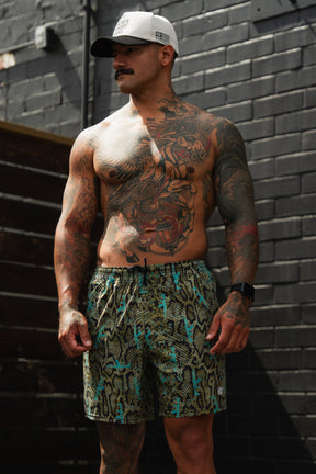 A muscular, tattooed man in a white cap and Tango Charlie Apparel Men's Performance Training Shorts | 5.5"–Lethal Venom stands shirtless against a dark brick wall, glancing to his left with a smartwatch on his wrist.