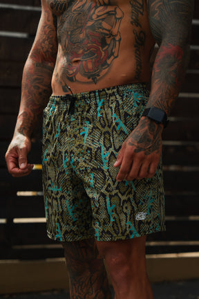 A heavily tattooed person wears Tango Charlie Apparel's Men's Performance Training Shorts | 5.5"- Lethal Venom in green and blue snake-print with a black smartwatch, standing before a dark wooden fence.