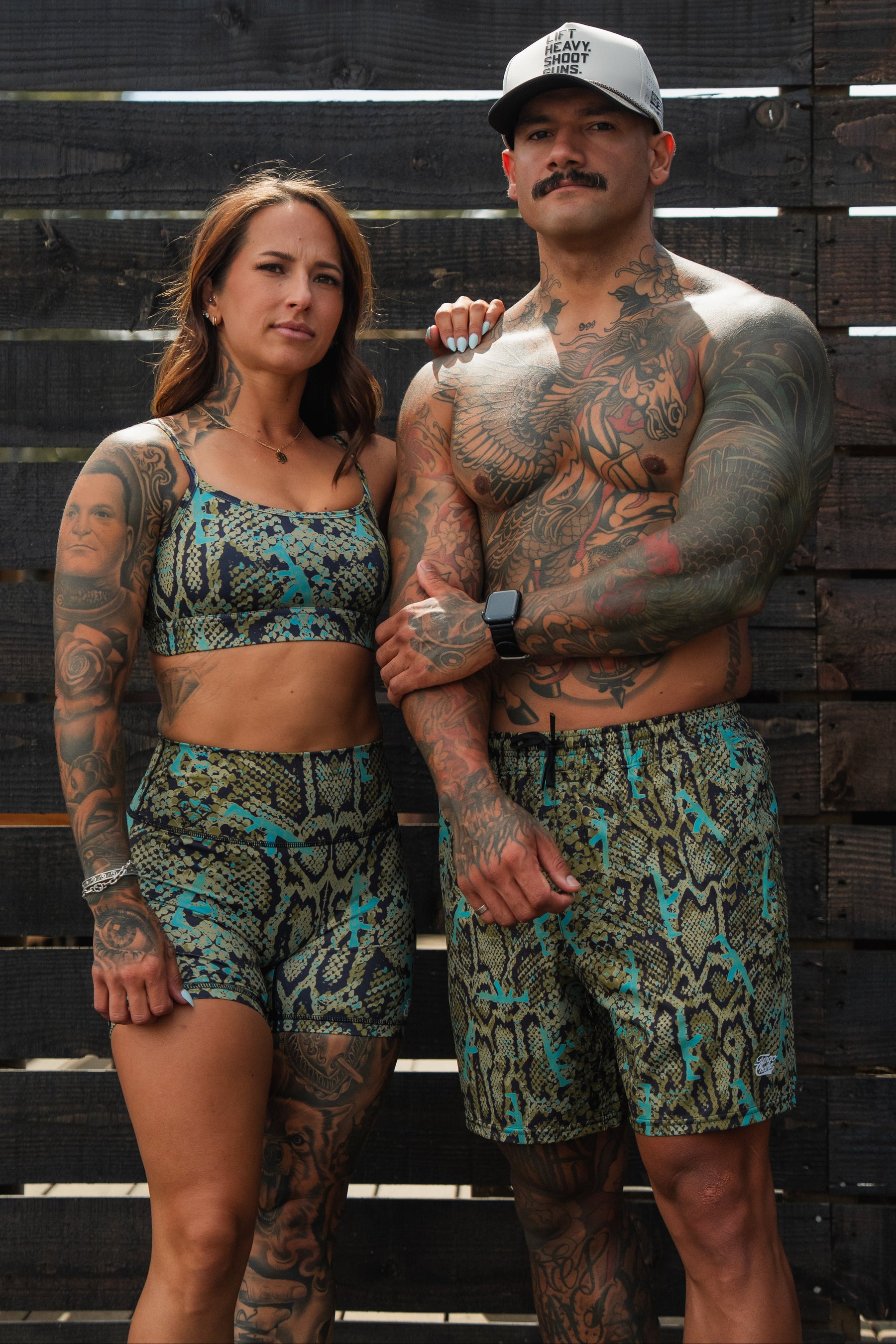 A tattooed woman and man pose confidently in matching snake-print outfits by Tango Charlie Apparel. She wears Women's High-Waisted Seamless Front Compression Training Shorts | Lethal Venom, while he stands shirtless in a hat against wooden planks.