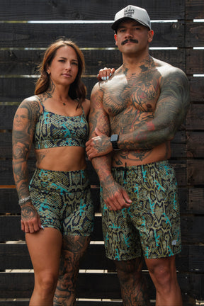 A tattooed woman and man pose confidently in matching snake-print outfits by Tango Charlie Apparel. She wears Women's High-Waisted Seamless Front Compression Training Shorts | Lethal Venom, while he stands shirtless in a hat against wooden planks.