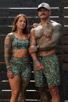 A tattooed man and woman stand together in eye-catching snake print activewear. The man, shirtless, wears Tango Charlie Apparel's Men's Performance Training Shorts | 5.5"- Lethal Venom—ideal hybrids for gym to swim—posing before a dark wooden fence.